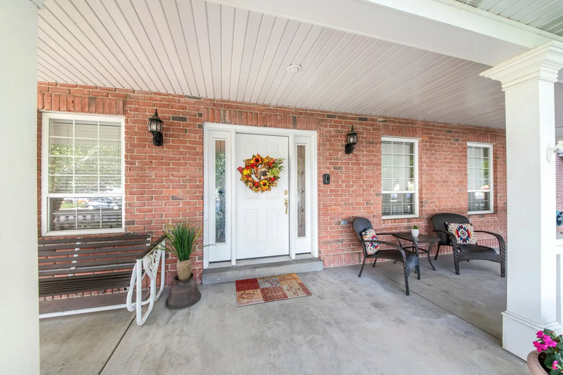 Covered brick front porch with a white door, wicker chairs, and a beadboard ceiling