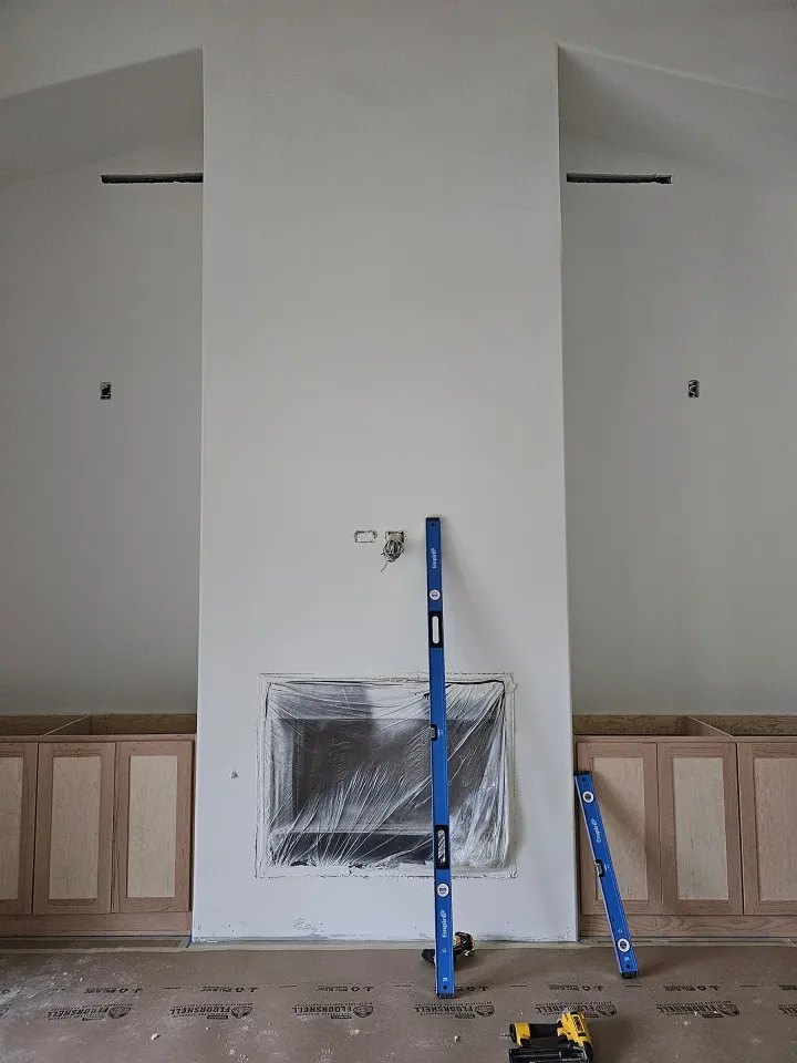 Freshly drywalled and mudded fireplace chase wall with the firebox opening masked in plastic, cut-in electrical boxes, two levels leaning against the wall, and floor protection paper down