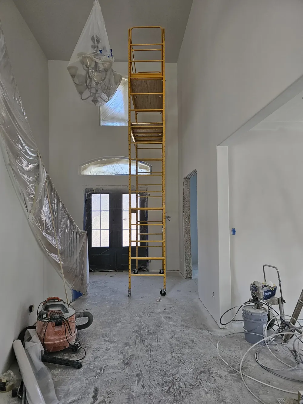 Two-story entry masked for painting, with a rolling scaffold tower, the chandelier bagged in plastic, the front door and stair rail sheeted off, a HEPA vacuum, and an airless sprayer